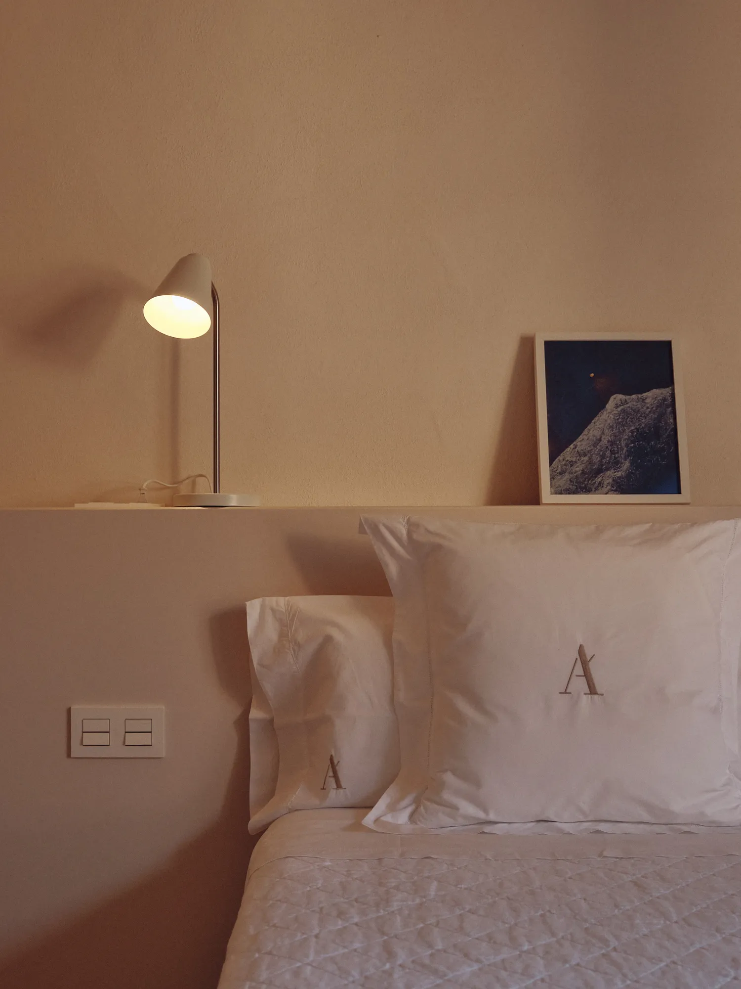 Rincón de dormitorio con cama vestida, almohadas con inicial A bordada, lámpara de lectura y cuadro abstracto.