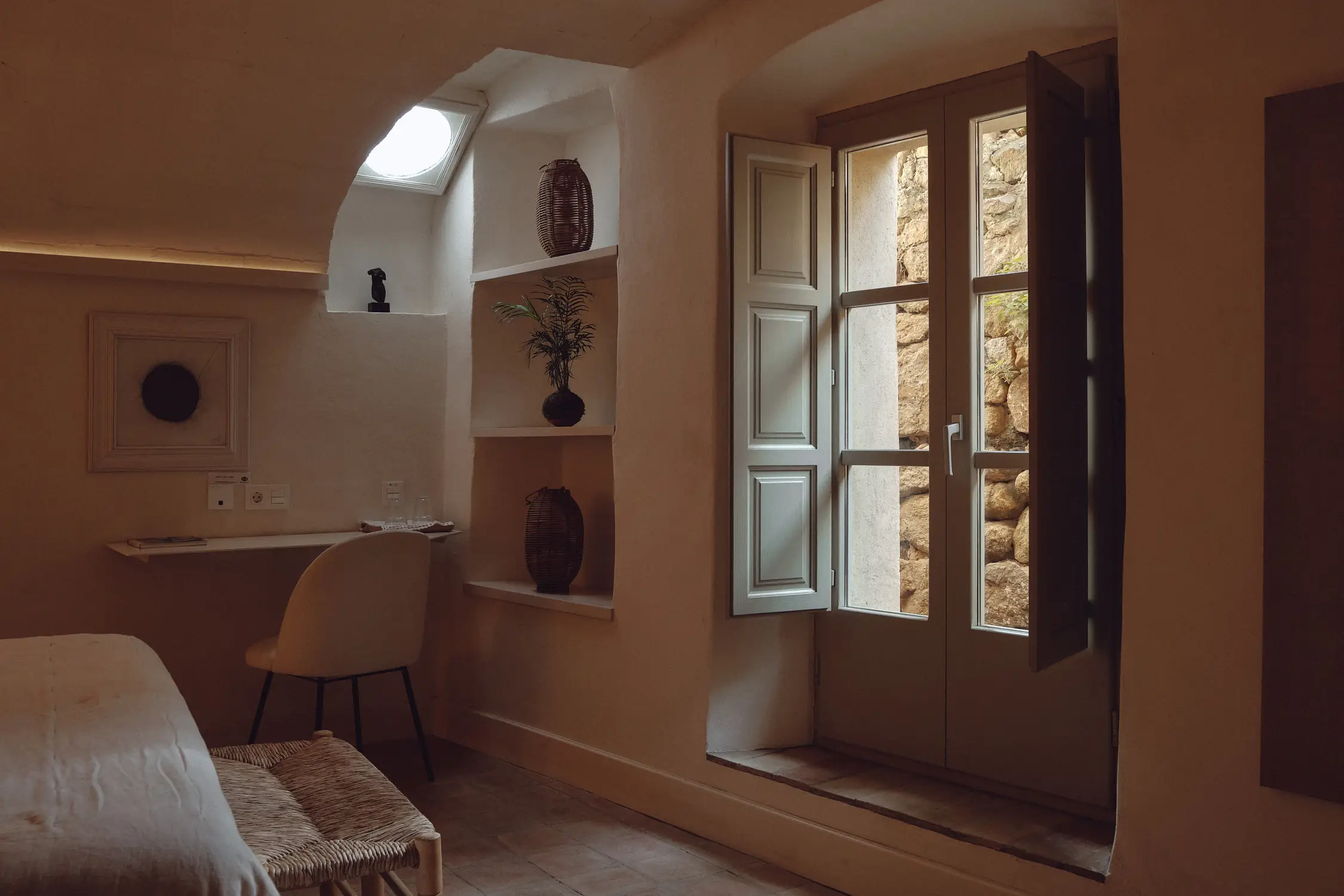 Acogedor interior mediterráneo con escritorio, luz natural cenital y ventana que abre a un muro de piedra.